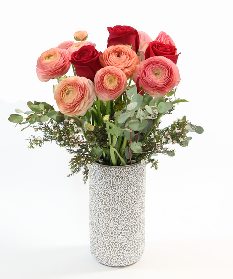 Soft ranunculus blooms accented with subtle seasonal touches, all arranged in a textured cylinder vase.